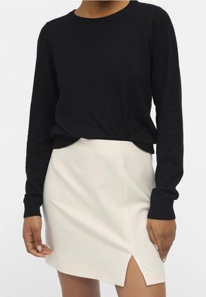 Vrouw die een zwarte top met lange mouwen draagt, ingestopt in een witte minirok met een voorkantssplit, staand tegen een effen witte achtergrond.