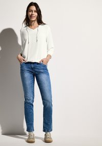 Lichtgewicht witte lange mouwen top met een trekkoord hals, gecombineerd met mid-blauwe slim-fit jeans en beige sneakers. Eenvoudige, casual outfit.