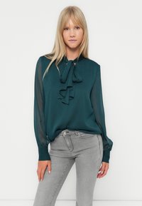 Τιρκουάζ μπλούζα με λεπτομέρεια φιόγκου στο λαιμό, μακριά διάφανα μανίκια και λείο υλικό· σε συνδυασμό με ανοιχτό γκρι εφαρμοστά τζιν.