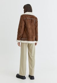 Chaqueta de ante marrón con cuello y puños de piel de oveja blanca, que presenta costuras horizontales y una cintura ceñida con cinturón, llevada sobre pantalones beige a rayas.