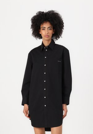 Schwarzes, oversized Hemdkleid aus Baumwolle mit durchgehender Knopfleiste, langen Ärmeln und einer Brusttasche mit dezentem Logo. Weiche Textur.