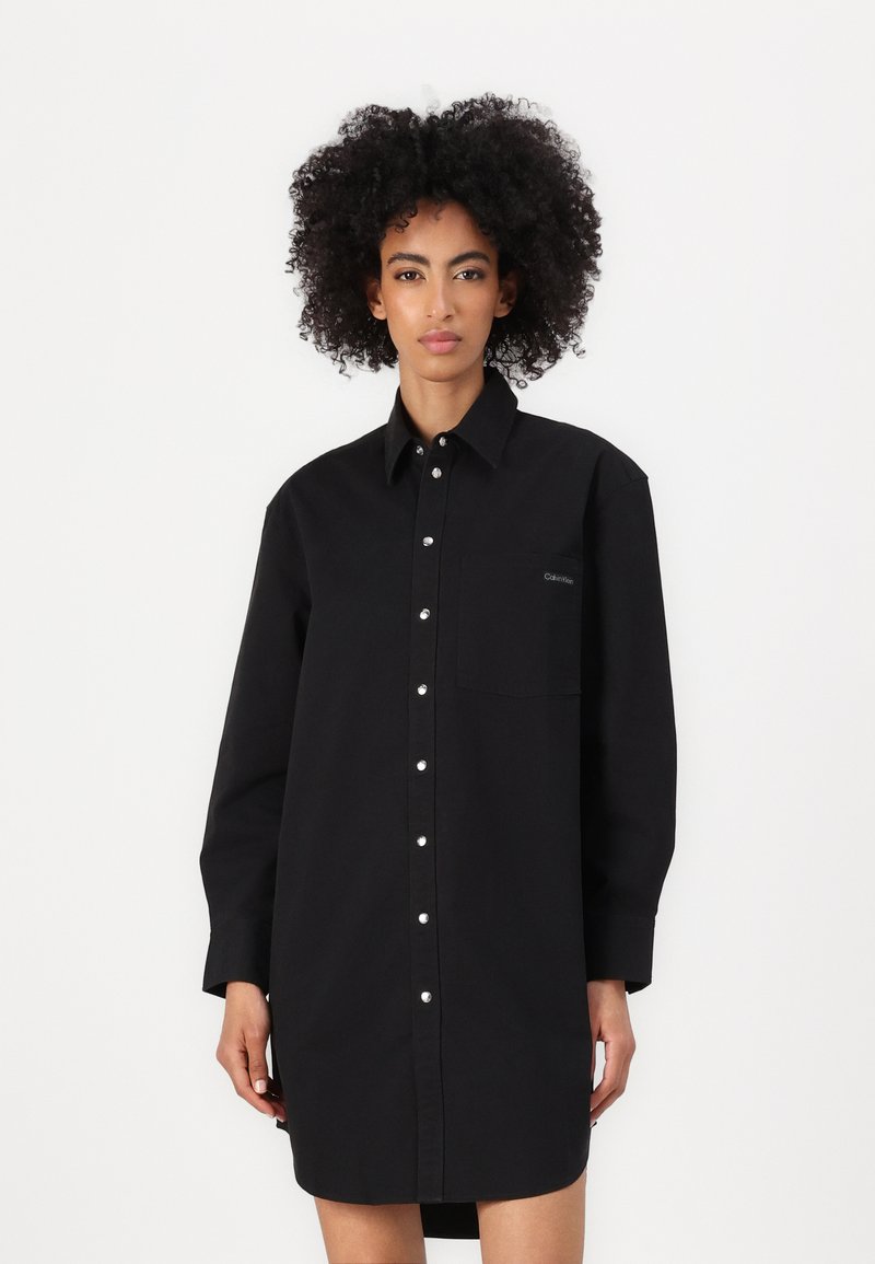 Abito camicia nero oversize in cotone, con chiusura a bottoni sul davanti, maniche lunghe e tasca sul petto con un logo discreto. Tessuto morbido.