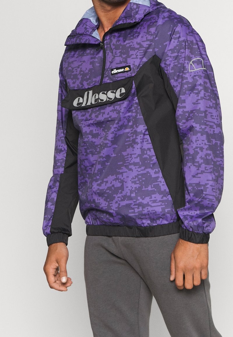 Lila och svart tröja med dragkedja, försedd med ett pixelmönster, elastiska muddarna och en logotypdekal framtill. Lättviktig tyg.