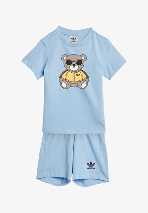 Jasnoniebieski strój Adidas dla malucha z krótkimi rękawami i szortami, na przedniej części koszulki z misiem w okularach przeciwsłonecznych i żółtej kurtce.