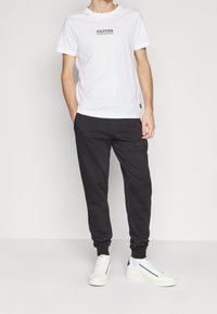 Mies, der ein weißes "Hilfiger" T-Shirt, schwarze Jogginghosen und weiße Sneakers trägt und vor einem hellen einfarbigen Hintergrund steht.