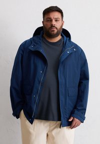 Marineblau leichte Jacke mit Kapuze, Frontreißverschluss und zwei Taschen. Verfügt über eine lockere Passform und strukturierten Stoff. Über einem dunklen Hemd getragen.