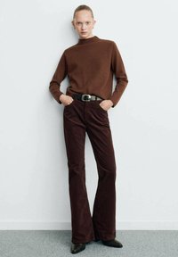 Pull en col roulé marron ajusté associé à un pantalon en velours côtelé bordeaux évasé. Ceinture noire avec une boucle en argent et chaussures noires pointues. Fond minimal.