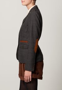 Personne portant un blazer gris à carreaux avec des coudières brunes et des accents bruns, associé à un short en velours côtelé marron et des collants noirs, se tenant de profil.