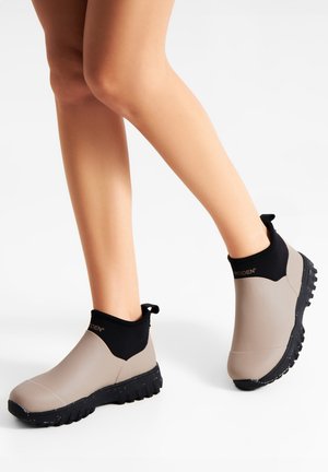 Laarzen met een slank ontwerp in lichtbeige en een zwarte, rekbare enkel. De zool is getextureerd en van rubber, wat grip en stabiliteit biedt.