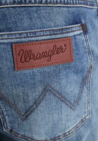 Primer plano de una tela de mezclilla que muestra un bolsillo trasero con una etiqueta de cuero marrón de Wrangler y costuras en zigzag con hilo naranja.