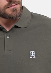 Polo verde oliva fabricado con tejido texturizado, que presenta un cierre de tres botones y un logo bordado en blanco en el lado izquierdo del pecho.