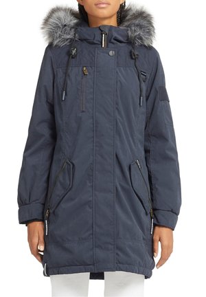 Femme portant un parka à capuche bleu marine avec une bordure en fausse fourrure grise, des poches zippées et des cordons, associée à un pantalon blanc.