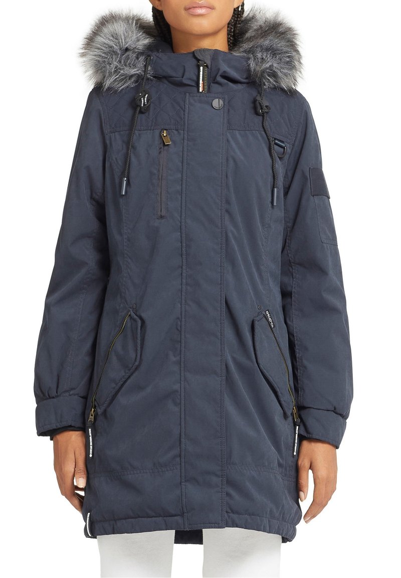 Femme portant un parka à capuche bleu marine avec une bordure en fausse fourrure grise, des poches zippées et des cordons, associée à un pantalon blanc.