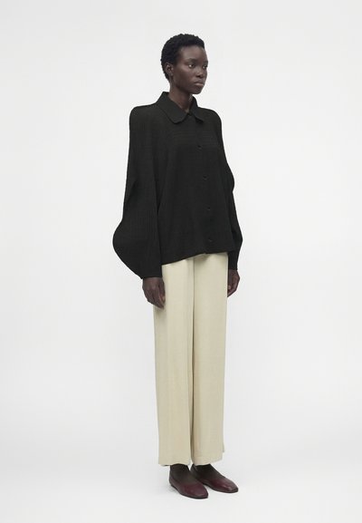 Γυναίκα που στέκεται φορώντας μια φαρδιά μαύρη πουκάμισα με κουμπιά και μακριά μπαλούν μανίκια, μπεζ παντελόνια με φαρδιά μπατζάκια και σκούρα καφέ παπούτσια σε λευκό φόντο.