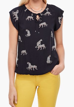 Mustahihaisen leopardikuosisen puseron, jossa nyöritys pääntiellä, yhdistetty keltaisiin housuihin, käsi taskussa.