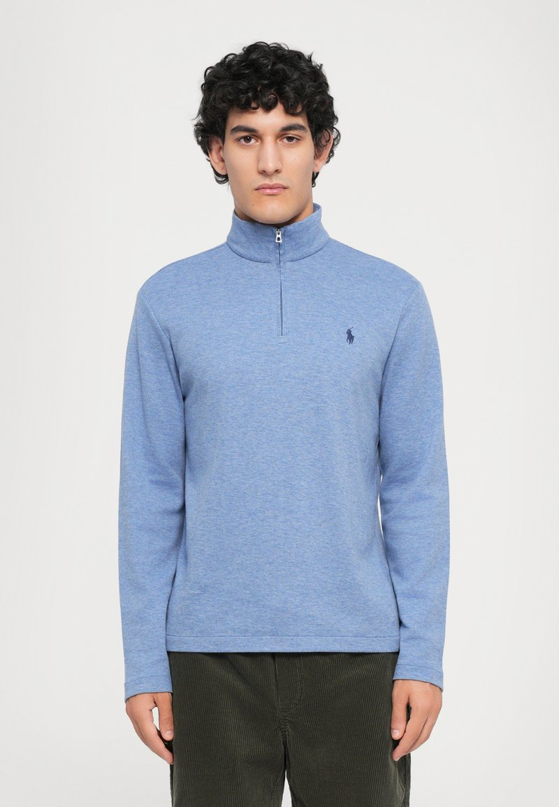 Pullover azul claro com gola de meia zíper, mangas longas e um pequeno logótipo escuro no lado esquerdo do peito. Tecido suave e texturizado, corte casual.