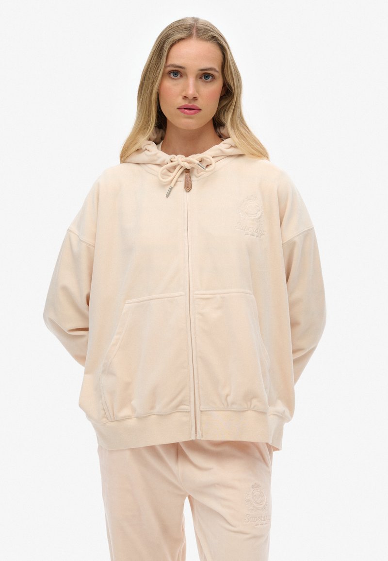 Haut à capuche zippé rose clair en matériau doux, présentant une capuche à cordon de serrage, des poches avant et un logo brodé sur la poitrine.