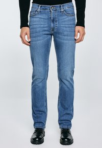 Mees, kes kannab kitsaste säärega siniseid teksapükse, musta pika varrukaga tuba ja musti nahast paeltega kingi, seisab ühevärvilise taustaga ees.