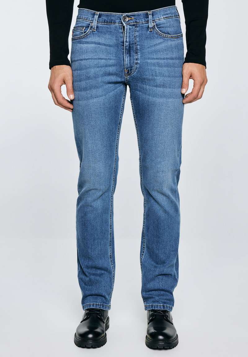 Mees, kes kannab kitsaste säärega siniseid teksapükse, musta pika varrukaga tuba ja musti nahast paeltega kingi, seisab ühevärvilise taustaga ees.