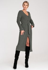 Langer, gerippter Strickcardigan in Dunkelgrün mit V-Ausschnitt, Frontknöpfen und einem Bindegürtel. Verfügt über einen schenkelhohen Schlitz und Glockenärmel.