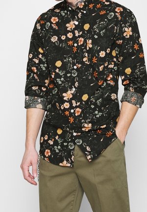 Mand iført sort blomsterprint skjorte med knappelukning og op rullede ærmer samt olivengrønne bukser, hånden i lommen, foran en ensfarvet baggrund.