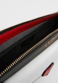 Pouch multi-scomparto in pelle nera e bianca con fodera rossa. Presenta una cerniera dorata e un logo impresso. Texture liscia e design strutturato.