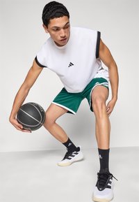Weißes ärmelloses Sportshirt mit schwarzen Akzenten, grüne Basketballshorts mit weißem Rand, schwarze Basketballschuhe, hält einen schwarzen Basketball.