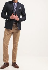Marineblauwe dubbelgeknoopte blazer met zilveren knopen, lichtblauw shirt, beige chinos en bruine leren schoenen. De pochet heeft een patroon.