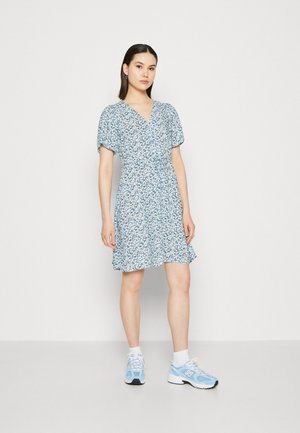 Robe portefeuille bleue à fleurs avec de courtes manches bouffantes et un ourlet froncé. Associée à des baskets bleu clair et des chaussettes blanches à la cheville.