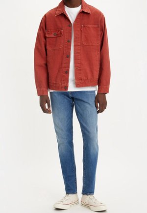 Chaqueta de mezclilla roja de algodón con cierre de botones, dos bolsillos en el pecho y un ajuste relajado, combinada con jeans azules y zapatillas blancas.