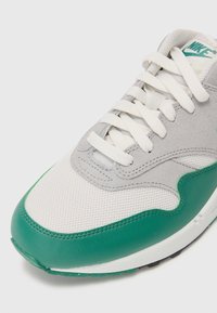 Sivé a biele tenisky s zelenými akcentmi, ktoré majú sieťovinový zvršok, polstrovaný golier a tradičné šnurovanie. Hladké a textúrované povrchy.