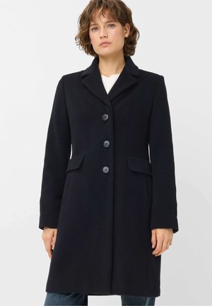 Woman wearing a dark navy knee-length coat with three buttons and flap pockets, standing against a plain white background.