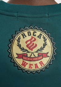 Türkiz pulóver, bézs színű, kör alakú hímzett logóval, amelyen a "ROCA WEAR" felirat és díszítőelemek, köztük láng és babérlevelek találhatók.