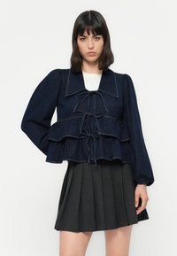 Donkerblauwe denim peplumjack met een grote kraag, poffende mouwen en trekkoordaccenten. Gecombineerd met een geplooide grijze rok.