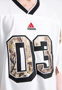 Balts sporta krekls ar melnu apdari, rotāts ar lieliem teksturētiem kamuflāžas skaitļiem "03" un izšūtu sarkanu Adidas logo uz pleca.
