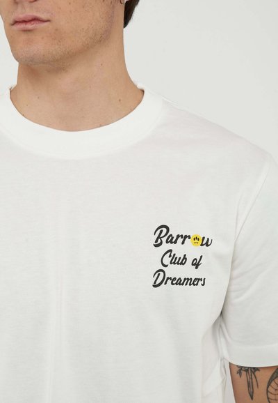 Giovane uomo che indossa una maglietta bianca con testo nero "Barrow Club of Dreamers" e una piccola faccina gialla sorridente vicino al petto, che mostra parte del braccio tatuato.