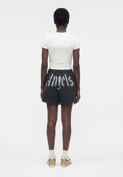 Άτομο στέκεται με την πλάτη, φοράει λευκό κοντομάνικο μπλουζάκι, μαύρες σορτς με το γράμμα "Angels" σε μεγάλο γοτθικό τύπο, λευκές κάλτσες και μπεζ αθλητικά παπούτσια.