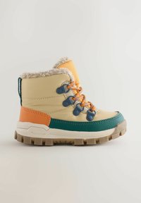 Bottine beige et verte avec un col en fourrure crème, des accents orange et bleu, une semelle en caoutchouc texturée et un système de laçage à œillets.