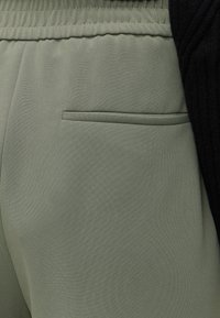 Gros plan sur un pantalon vert à taille élastique montrant une poche arrière horizontale et une partie d'un pull noir côtelé.