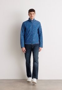Casaco azul estampado com colarinho alto, fecho éclair à frente e mangas compridas, usado sobre calças de ganga escuras e ténis brancos. Textura suave.