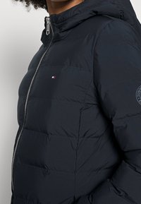 Marineblå pufferjakke med lynlås, hætte, quiltet tekstur og et lille logo på brystet. Har elastiske manchetter og forneden.