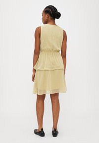 Rochie fără mâneci, galben deschis, cu un design în straturi și un model discret de buline. Prezentă o talie elastică și o margine până la genunchi.