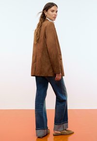 Blazer en suede marron avec une coupe décontractée, associé à un jean flare bleu avec des ourlets retroussés et des chaussures slip-on olive. Fond clair.