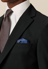 Schwarzer Anzug aus strukturiertem Stoff, weißes Hemd und eine braune gemusterte Krawatte. Ein blauer Einstecktuch im oberen Tascheneck setzt einen Farbakzent.
