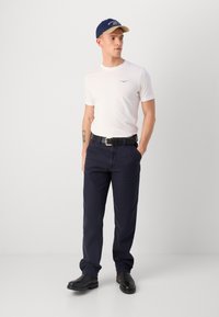 Witte T-shirt met korte mouwen en logo, marineblauwe broek, zwarte riem en zwarte schoenen. Blauwe pet met crème rand. Eenvoudige, casual outfit.