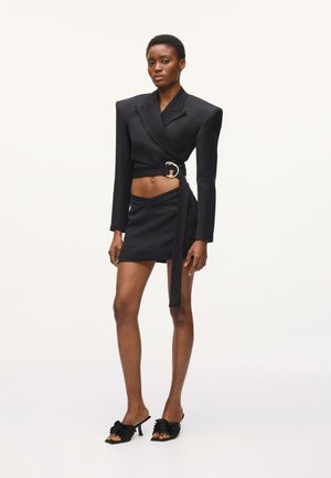 Mujer vistiendo un blazer corto negro estructurado con un cinturón ancho, una minifalda asimétrica a juego y sandalias negras de tacón con volantes.