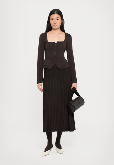 Brown fitted cardigan with square neckline and six buttons, paired with a pleated long skirt. Model holds a black cylindrical bag.