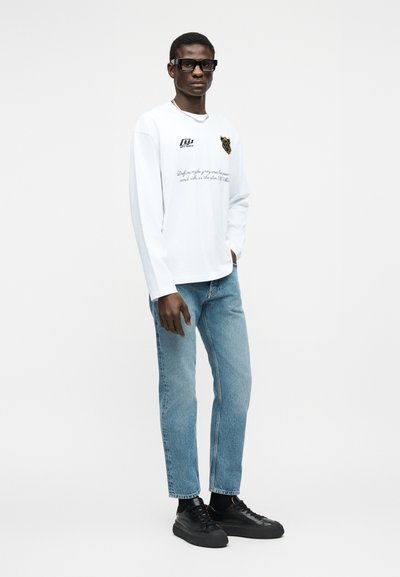 Νεαρός άνδρας με μαύρα γυαλιά, λευκό πουκάμισο με μακριά μανίκια και λογότυπα, ανοιχτό μπλε τζιν, μαύρα αθλητικά παπούτσια, στέκεται μπροστά σε λευκό, απλό φόντο.