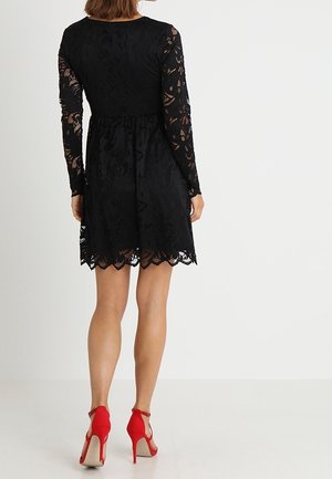 Femme portant une robe noire en dentelle longueur genoux à manches longues et des chaussures rouges à talons hauts avec bride à la cheville, se tenant devant un fond blanc uni.