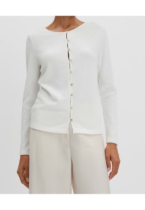 Kvinde iført en hvid tekstureret cardigan med knapper og lange ærmer, kombineret med løse, lysebeige bukser med vide ben mod en ensfarvet baggrund.